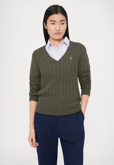 Olivovo zelený pletený sveter s vrkočovým vzorom, výstrihom do V, rebrovaným spodným lemom a manžetami, s malým oranžovým logom na ľavej strane hrude. Nosený cez svetlofialovú košeľu s golierom.