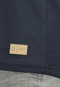 Navyblaues T-Shirt mit strukturierter Oberfläche, versehen mit einem beigen rechteckigen Etikett, auf dem das „ISOLID“-Branding näher am Saum gestickt ist.