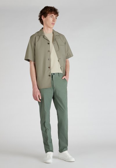 Chemise décontractée à manches courtes de couleur vert olive avec des boutons marron, associée à un pantalon à motifs vert clair et à des baskets blanches. Design minimaliste.