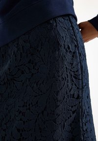 Mørkeblå blonde nederdel med blomsterprint, struktureret overflade og en tætsiddende talje, delvist dækket af en mørkeblå top.