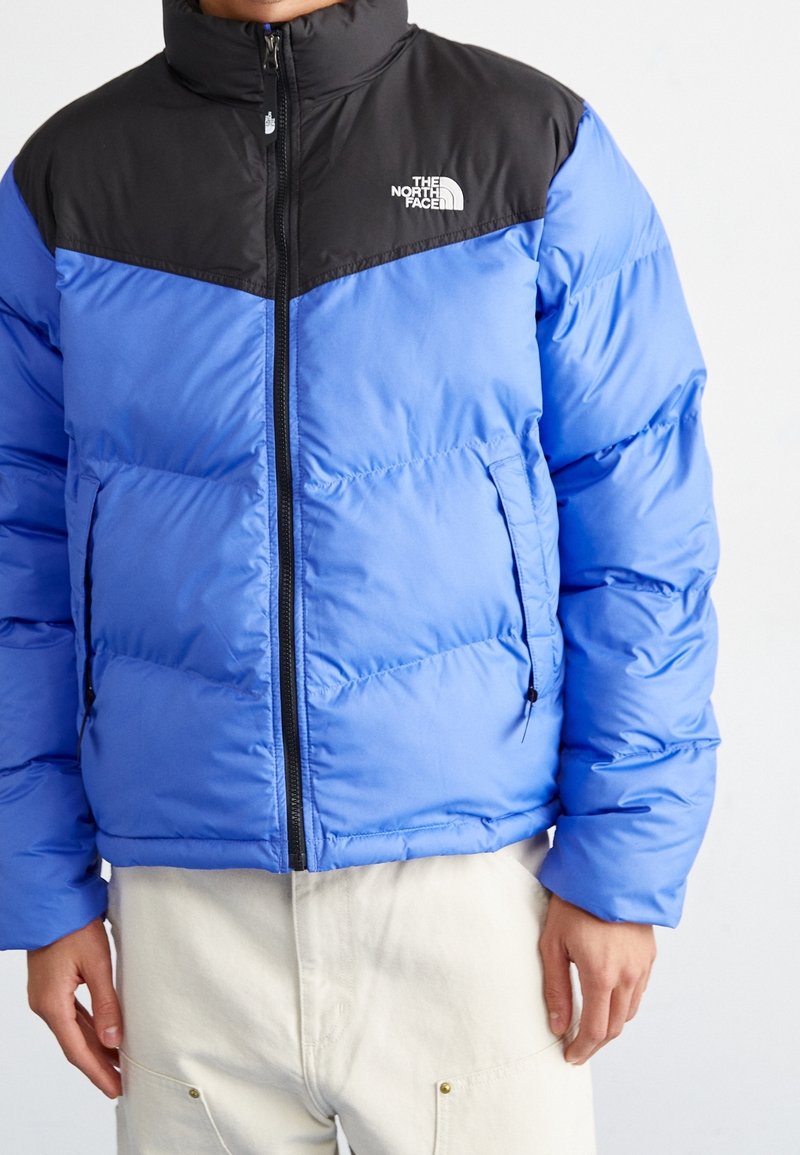 Veste matelassée bleue avec un panneau noir sur l'épaule, fermeture éclair sur le devant et deux poches latérales. Présente un rembourrage piqué et un logo sur la poitrine.