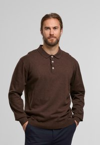 Mann mit Bart und zurückgebundenem Haar, der ein braunes Langarm-Polohemd und eine marineblaue Hose trägt, steht mit den Händen in den Taschen vor einem grauen Hintergrund.