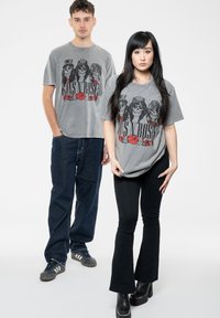 Gray graphic t-shirts featuring the text "Guns N' Roses" and three skulls with roses. Models wearing dark pants and shoes.