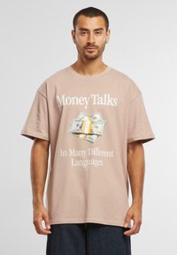 Tricou din bumbac bej cu mânecă scurtă, cu grafică "Money Talks" și ilustrație cu bancnote de dolari americani, având text alb îndrăzneț dedesubt.