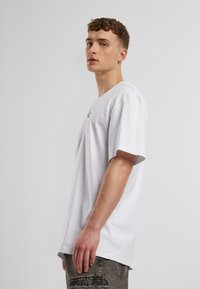 T-shirt en coton blanc avec un col rond et des manches courtes, arborant un petit logo noir sur le devant. Porté avec un pantalon cargo gris.