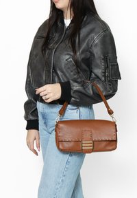 Borsa a spalla in pelle marrone con texture a granuli, chiusura in hardware dorato e tracolla rimovibile, indossata da una persona in bomber grigio e jeans chiari.