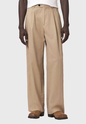 Mężczyzna w beżowych szerokich spodniach, białym topie i brązowych, teksturowanych butach, stoi na tle jednolitego jasnego tła.