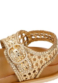 Sandal in oro intrecciato con una cinghia intrecciata e dettagli metallici, dotato di una suola piatta color tan e di una fibbia rettangolare per la regolazione.