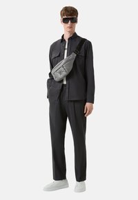 Zwarte button-up shirt, grijze puffer heuptas, op maat gemaakte donkere broek en witte sneakers. Model draagt oversized zwarte zonnebril.