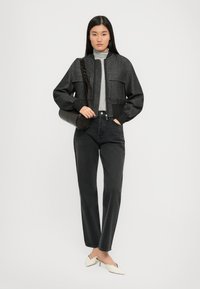 Veste bomber grise avec poches avant, col roulé gris clair, jeans foncés et mules blanches à talons. Sac à bandoulière noir avec une sangle.