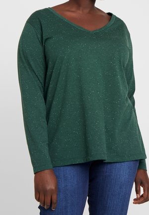 Femme portant un pull vert à manches longues avec col en V et taches blanches, associé à un jean bleu, vue du cou jusqu'aux hanches.