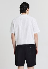 Mann mit dunklem, gewelltem Haar trägt ein weißes Kurzarm-Hemd mit Streifen, das in schwarze Shorts mit Knopftaschen gesteckt ist, und steht vor einem schlichten Hintergrund.