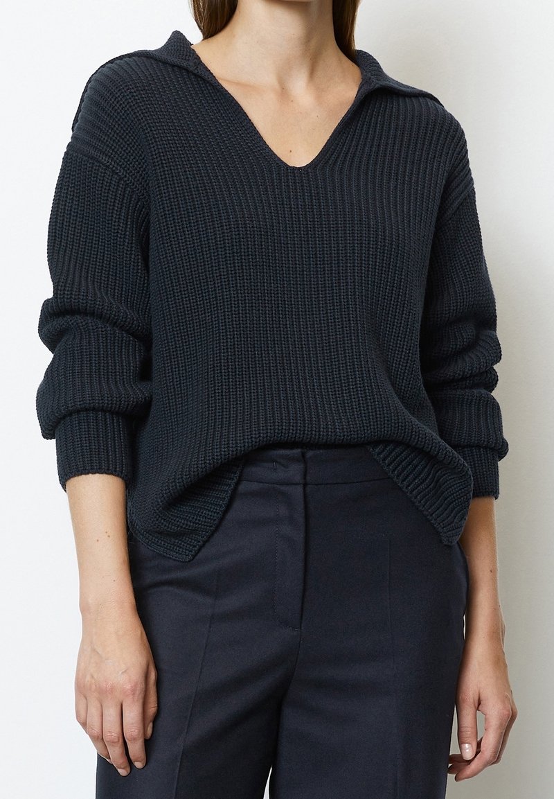 Femme portant un pull en tricot côtelé bleu marine foncé avec un col en V et un pantalon noir ajusté, les mains relâchées le long du corps.