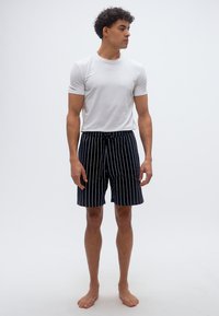 Άνδρας που στέκεται ξυπόλυτος, φορώντας μια απλή λευκή κοντομάνικη μπλούζα και μαύρο σορτς με κάθετες ρίγες, μπροστά σε αδιάφορο λευκό φόντο.