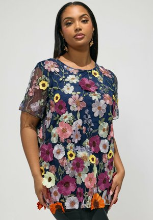 Donna che indossa una maglia trasparente blu navy con motivi floreali multicolori ricamati, in piedi davanti a uno sfondo chiaro e uniforme.