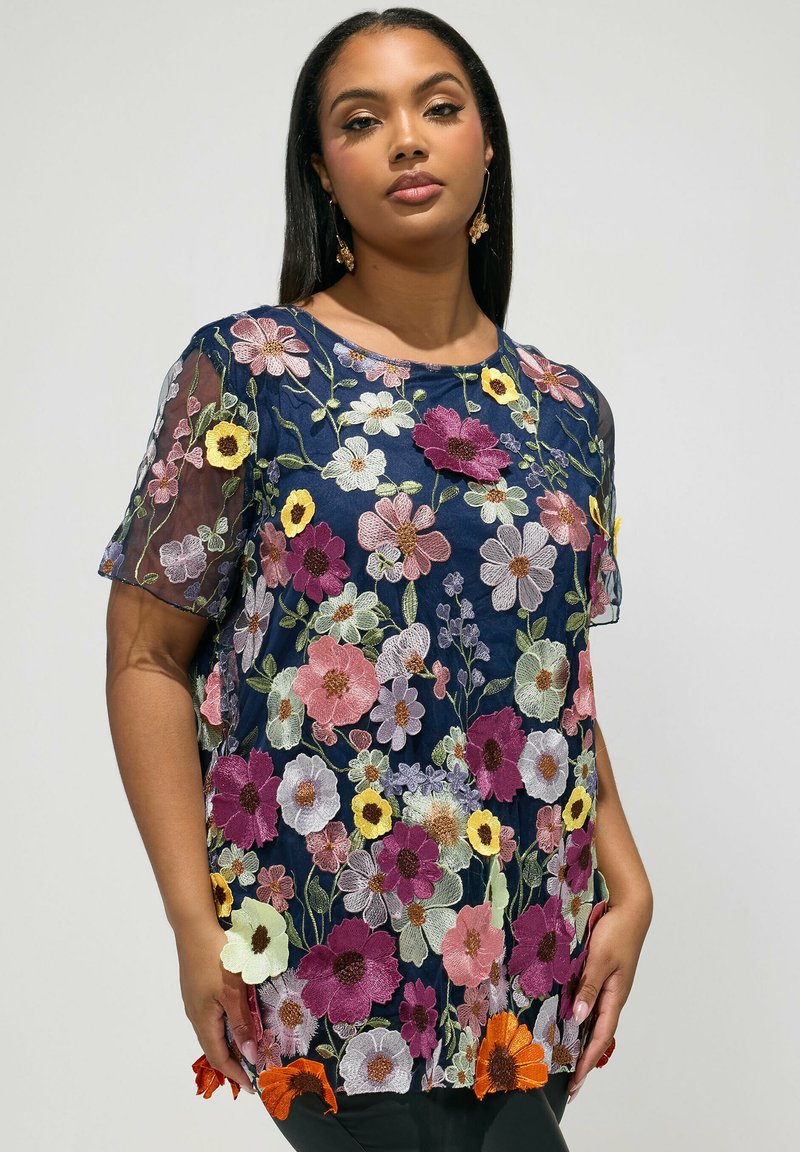 Femme portant un haut bleu marine transparent avec des motifs floraux multicolores brodés, debout devant un fond clair uni.