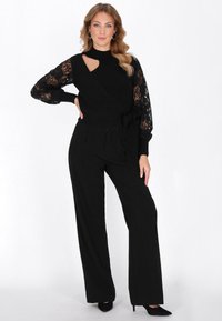 Czarny dzianinowy top z koronkowymi rękawami, z wycięciem, w połączeniu z czarnymi, wysokopasażowymi spodniami w szerokim kroju i czarnymi szpilkami. Gładka tekstura w całej stylizacji.