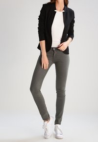 Černý blazer s puntíky přes bílou košili, doplněný o přiléhavé šedé džíny a bílé tenisky. Blazer má límcový design a texturovanou látku.