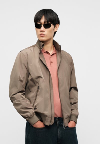 Νεαρός άνδρας με μαύρα γυαλιά ηλίου, τζάκετ ταμπά με φερμουάρ πάνω από ένα πουκάμισο πόλο σε σκόνη ροζ, με τα χέρια στις τσέπες μαύρων τζιν, ουδέτερο φόντο.