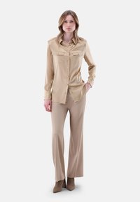 Camicia beige in satin con maniche lunghe e due tasche sul petto, abbinata a dei pantaloni beige. L'outfit presenta un design aderente e una texture liscia.