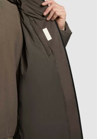 Interior de chaqueta con cremallera de color marrón oscuro, con textura suave, que cuenta con un bolsillo para la etiqueta y un forro ligeramente acolchado para aislamiento.