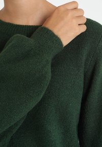 Pull en maille vert foncé avec un large col et un tissu texturé. Dispose de longues manches amples et d'une apparence douce et confortable.