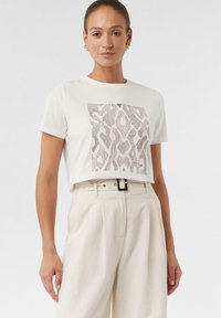 Biała koszulka z teksturowanym brązowym wzorem z przodu. Połączona z beżowymi spodniami w wysokim stanie i czarnym paskiem.