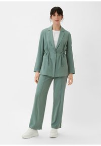 Lichtgroen pakset met een op maat gemaakte blazer met trekkoorddetails in de taille en wijduitlopende broeken, gecombineerd met witte sneakers.
