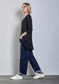 Svart långärmad topp med sidoklyftor, kombinerad med mörkblå vida jeans. Vita sneakers med ljusgrå detaljer. Enkel, avslappnad look.