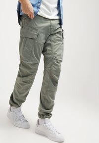 Olivgrön cargobyxor med flera stora fickor, smalnande muddar, mjukt material och en avslappnad passform, tillsammans med vita sneakers.