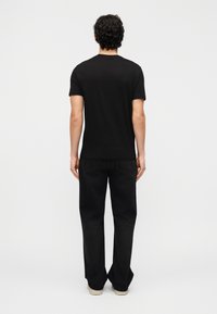 Άνδρας με σγουρά μαλλιά στέκεται με την πλάτη προς τα εμπρός, φοράει απλό μαύρο μπλουζάκι, μαύρο τζιν και ανοιχτόχρωμα παπούτσια μπροστά σε λευκό φόντο.