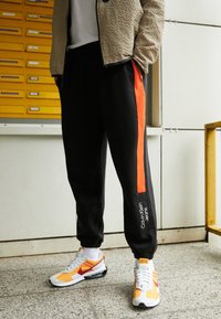 Black sweatpants with an orange side stripe and logo; paired with white and yellow sneakers. Beige textured jacket and plain white shirt.