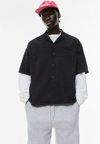 Cămașă din bumbac neagră, cu mâneci scurte și închidere cu nasturi, având un logo subtil. Pur tată peste o cămașă albă cu mâneci lungi, asociată cu pantaloni de trening gri.