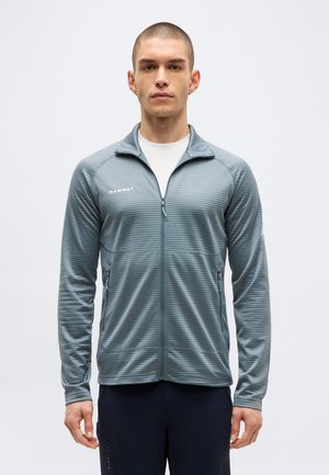 Mees, kes kannab helesinist tõmblukuga tekstuuriga jopet „Mammut“ logoga rinnal ja tumedaid pükse, seisab ühevärvilise tausta ees.