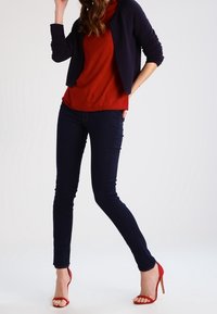 Červený top s žebrovaným vzorem pod tmavě modrým kardiganem, v kombinaci s tmavě modrými přiléhavými džínami a červenými sandály na vysokém podpatku, stojící na světlém pozadí.