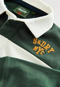 Green and white striped polo shirt with a white collar, featuring embroidered "S&DRY NYC" in orange. Made of textured cotton fabric.