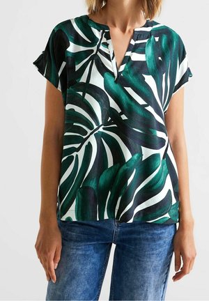 Vrouw die een blouse met korte mouwen draagt met een tropische bladprint in groen en zwart, en een blauwe spijkerbroek, staand tegen een effen achtergrond.