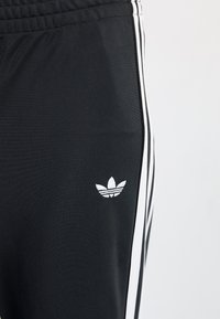 Black athletic pants made of smooth fabric, featuring white side stripes and a small white logo on the left leg. Elastic waistband included.