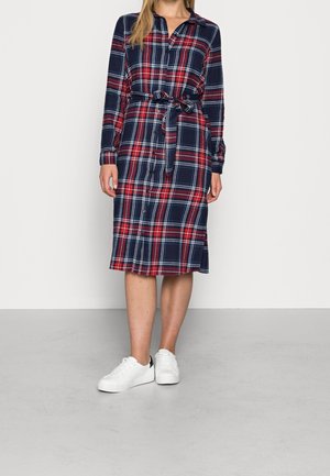 Kvinde iført en knælængde skjortekjole i marineblå, rød og hvid tern med bindebælte og hvide sneakers, stående på gråt gulv.