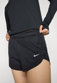 Musta urheilushortsit, joissa on joustava vyötärönauha, sivutaskut ja hopeanvärinen Nike-logo helmassa. Kevyt ja hengittävä kangas.