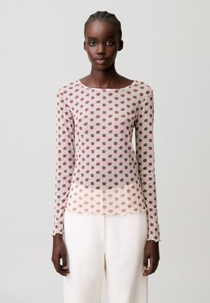 Joven mujer que lleva una blusa floral transparente de manga larga con bordes ondulados y pantalones blancos de talle alto, de pie contra un fondo liso.