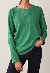Sweat-shirt vert en maille gaufrée avec une coupe décontractée, un col en V et des manches longues, associé à un jean foncé. Texture lisse et couleur vibrante.