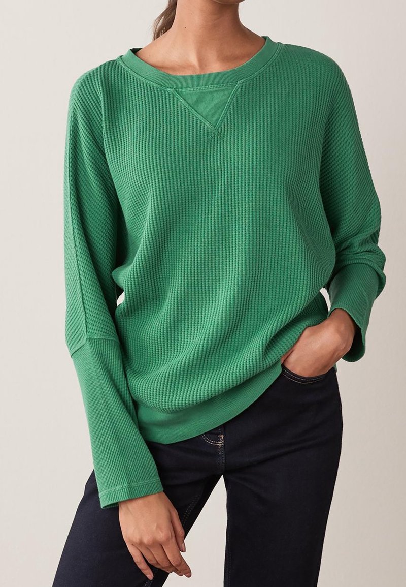 Sweat-shirt vert en maille gaufrée avec une coupe décontractée, un col en V et des manches longues, associé à un jean foncé. Texture lisse et couleur vibrante.