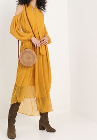 Żółta sukienka bez ramiączek, z długim rękawem, wykonana z lekkiej tkaniny, z detalem w postaci związania. Połączona z brązowymi kozakami za kolano i okrągłą plecioną torbą.