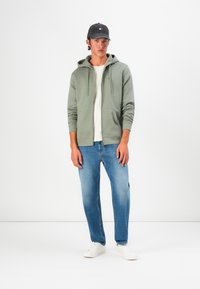 Joven vestido con una sudadera gris con cremallera, camiseta blanca, jeans azules, zapatillas blancas y gorra de béisbol negra, de pie contra un fondo blanco.
