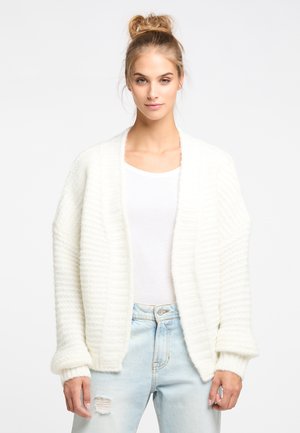 Weißer, strukturierter Cardigan mit lockerer Passform und weiten Ärmeln, getragen über einem eng anliegenden weißen Top und hellblauen, verwaschenen Jeans.