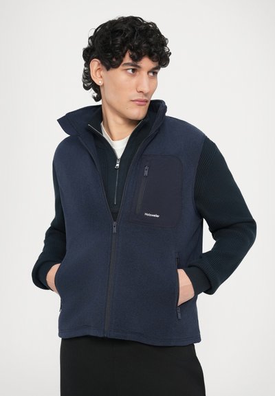 Jachetă din fleece, culise cu fermoar, de culoare bleumarin, cu mâneci din tricot cu aspect texturat și nervuri, prevăzută cu buzunar pe piept și buzunare laterale cu fermoar.