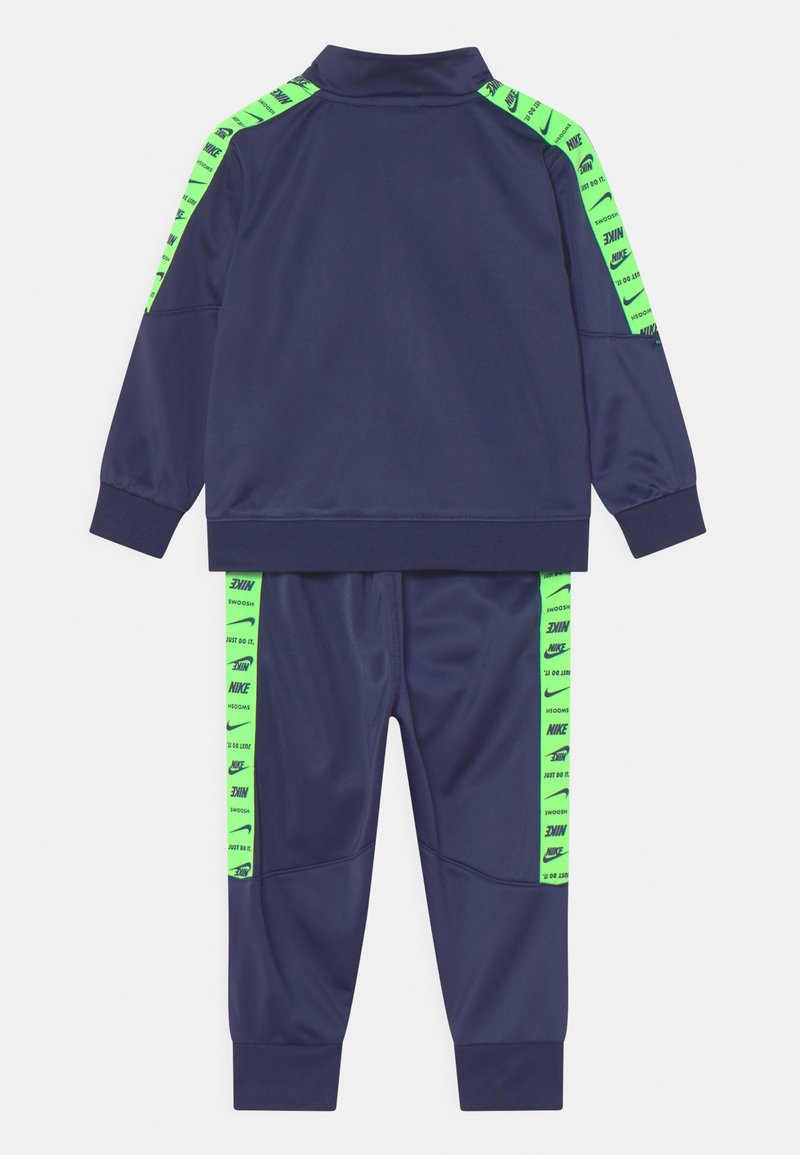 Chándal azul marino con acentos verdes de marca en los hombros y pantalones. Presenta un corte atlético, cuello alto y puños acanalados.