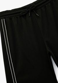 Zwarte joggers van een gestructureerde stof, met een elastische tailleband en een trekkoord, en twee witte verticale strepen langs de zijkanten.