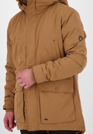 Mand iført beige hættejakke til vinter med klaplommer og synligt logo på venstre ærme og nederste lomme.